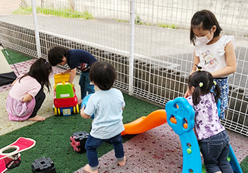 5人の子供がフェンスのある広場で滑り台や、車の押しぐるまなどで遊んでいる